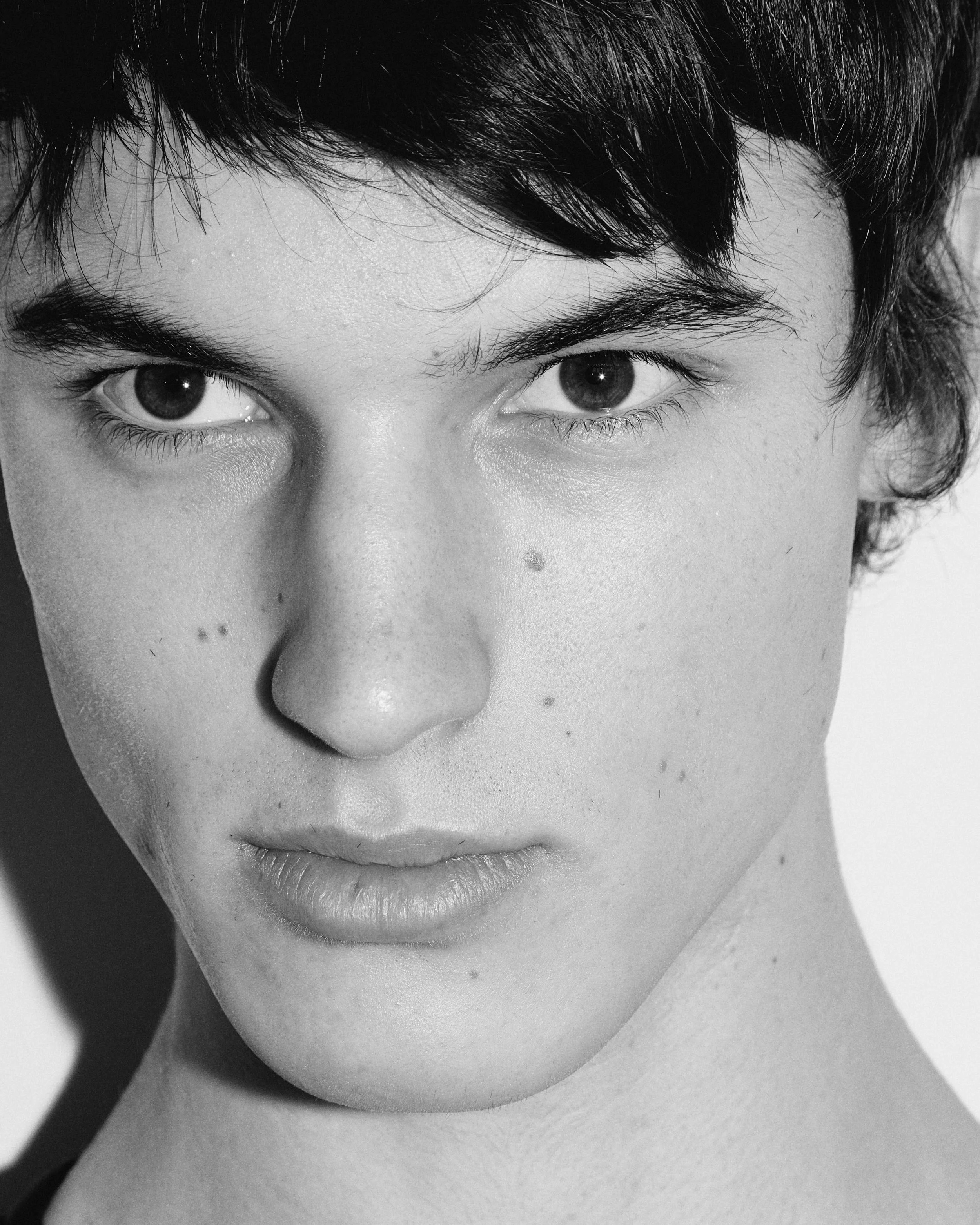 Close-up black and white portrait of a young person's face with short dark hair, intense eyes, and minimal makeup.