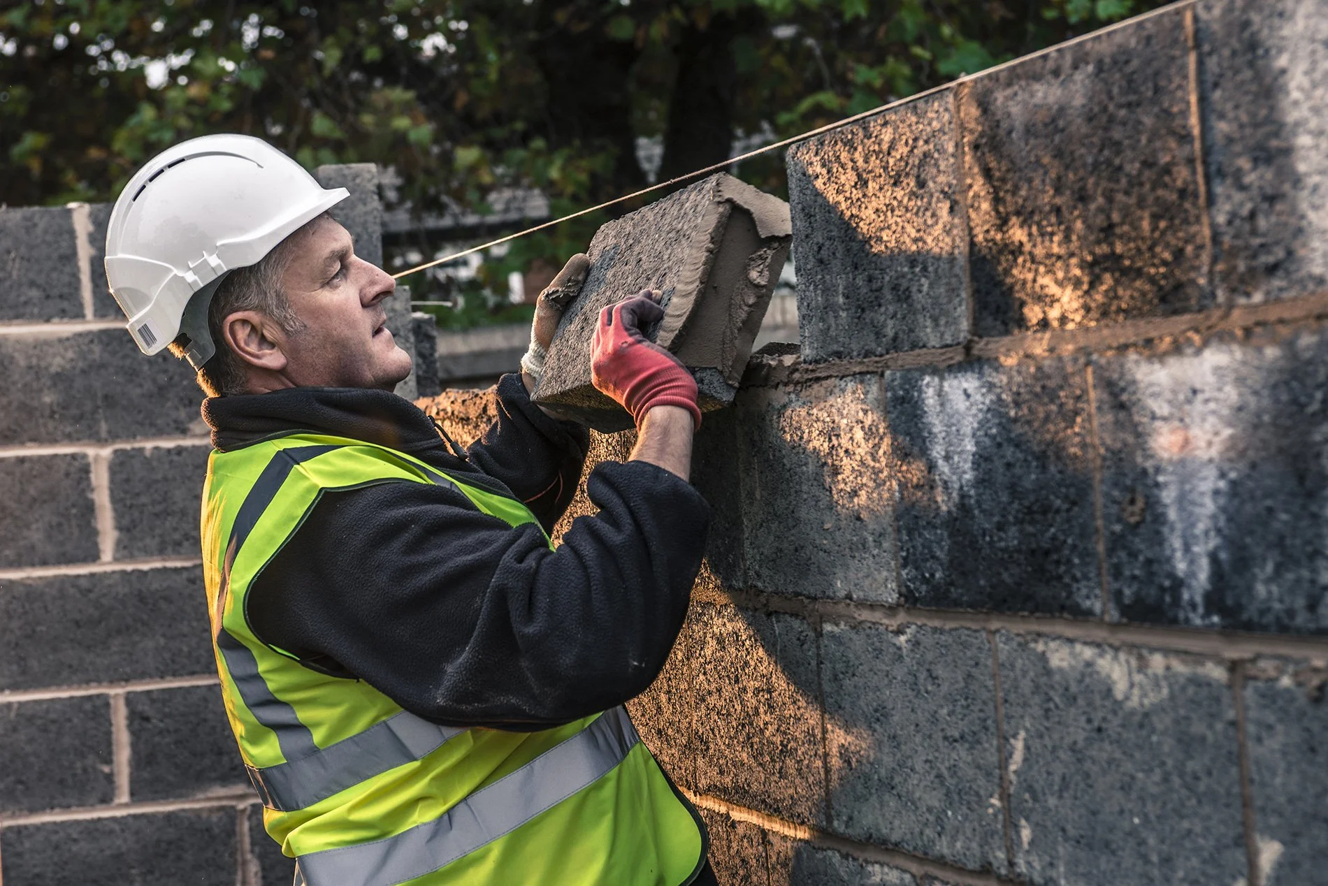 A construction worker wearing a white safety helmet, black hoodie, and a yellow reflective vest stacking concrete blocks on a wall.