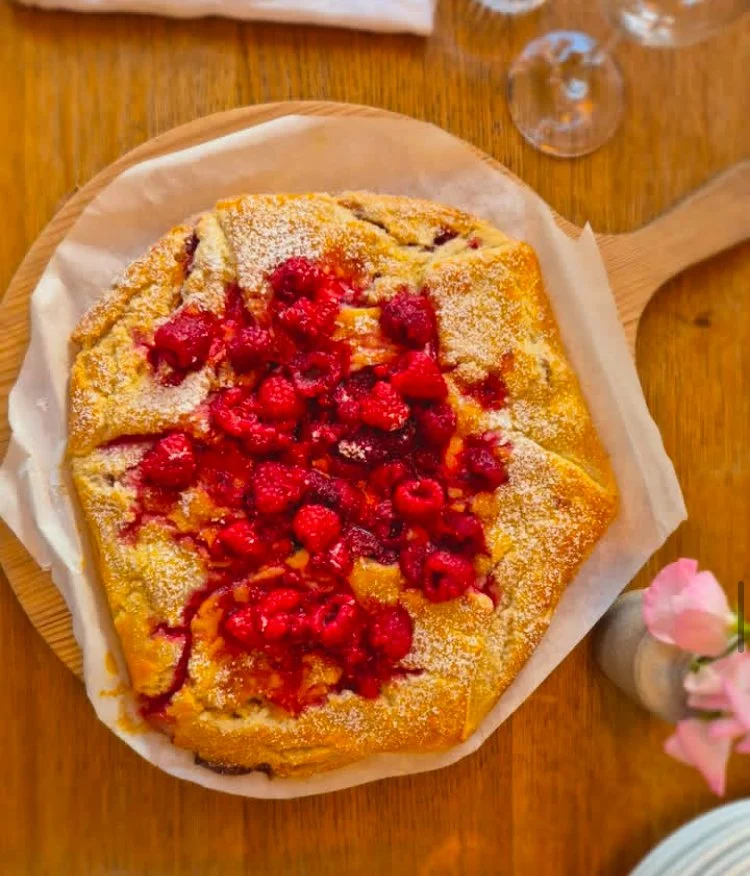 Berry-topped dessert 或 pastry on a wooden serving tray, dusted with powdered sugar, on a wooden table with a flower in a vase and glasses in the background.