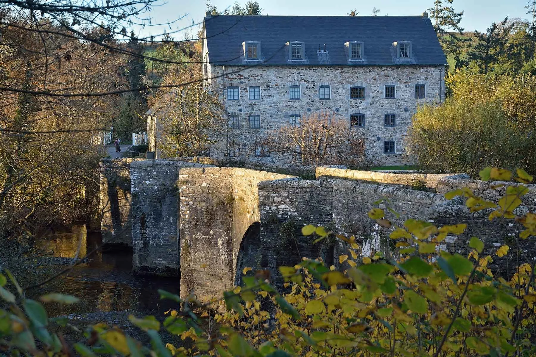 Stone bridge over a river in front of a large stone building with multiple windows, surrounded by trees with autumn foliage.