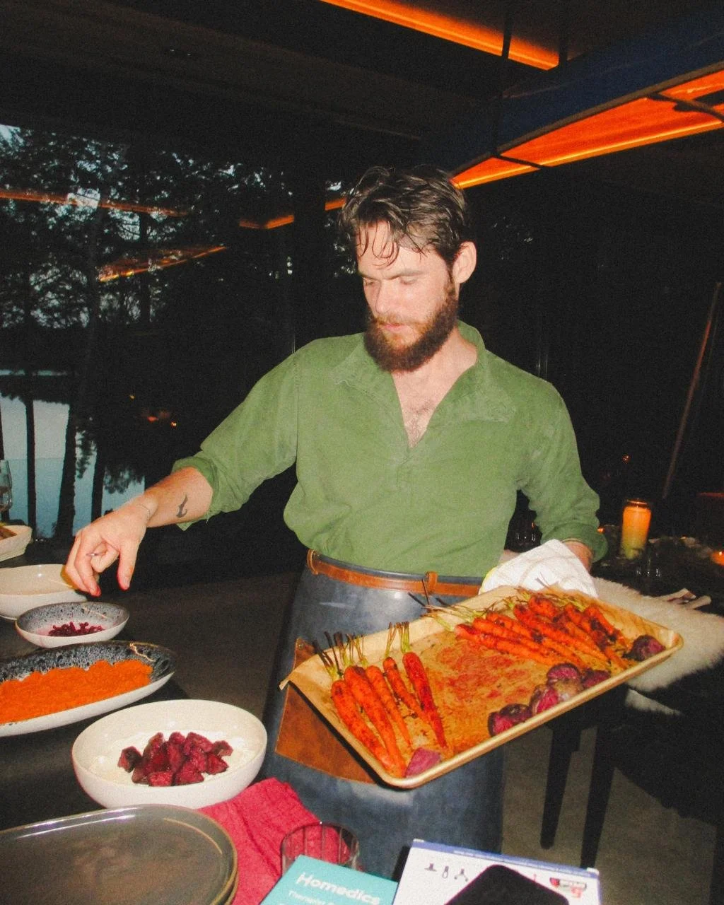 A man with a beard in a green shirt prepares food at a restaurant or bar with a lake view and trees in the background, under an orange-lit ceiling.