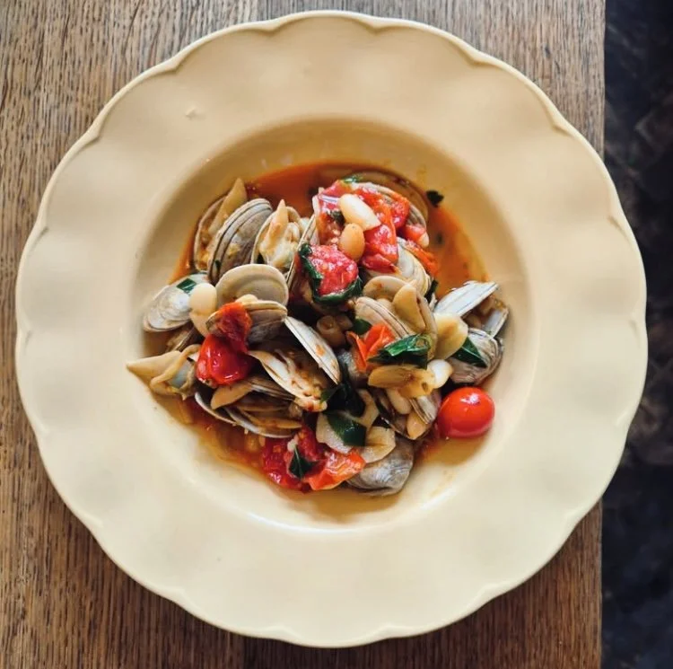 Seafood dish with clams, cherry tomatoes, herbs, and sauce on a cream-colored plate.