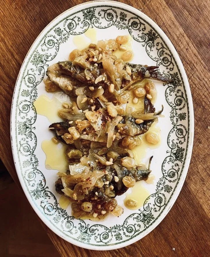 A white oval plate with decorative green floral border holding cooked small fish topped with chopped garlic, served with olive oil, on a wooden table.