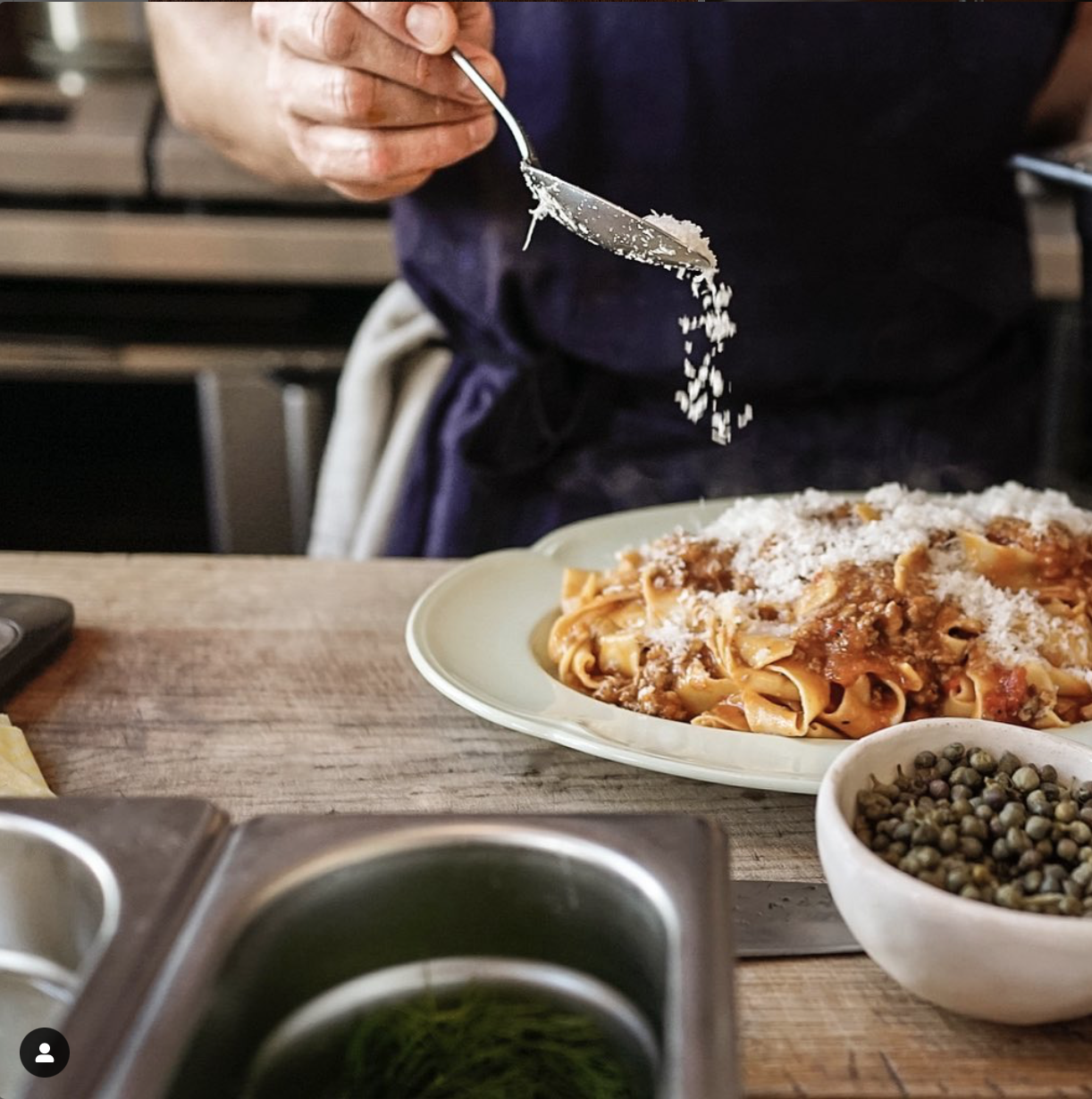 A person sprinkling grated cheese onto a plate of pasta with meat sauce, with a bowl of green peppercorns nearby, in a kitchen setting.