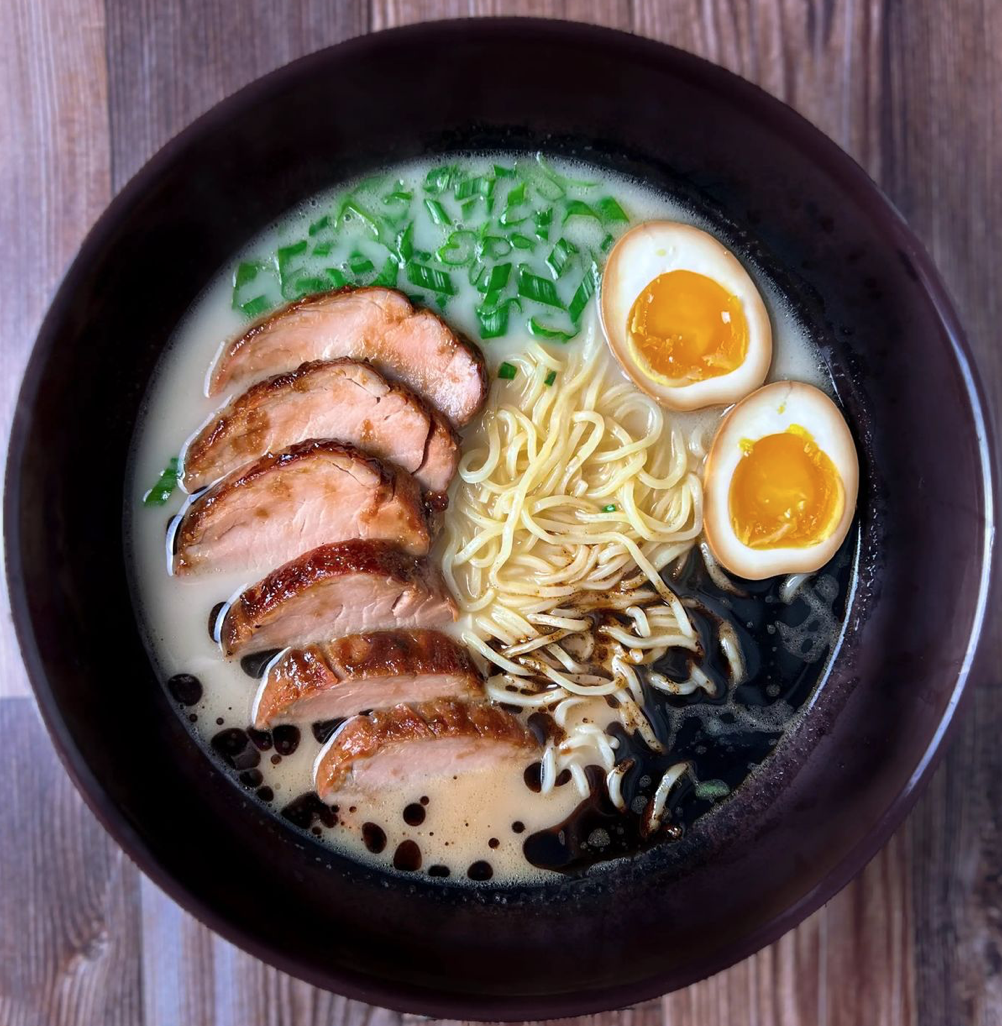 A bowl of ramen with sliced pork, soft boiled eggs, chopped green onions, noodles, and a creamy broth, served on a wooden table.