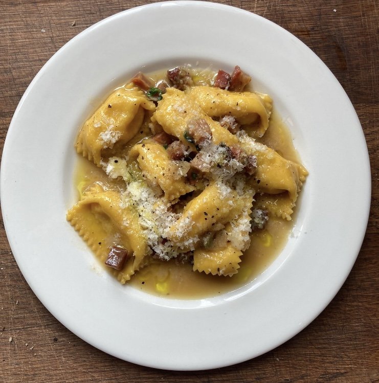 Plate of stuffed pasta, likely pasta shells, topped with cheese and served with a light broth and bits of meat, possibly bacon or ham.