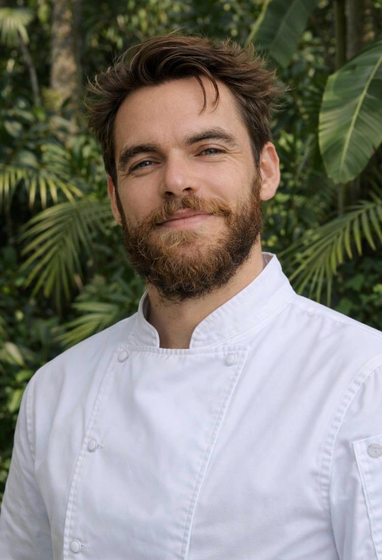 Portrait of a man with a beard in a white chef's coat, standing outdoors with green foliage in the background.