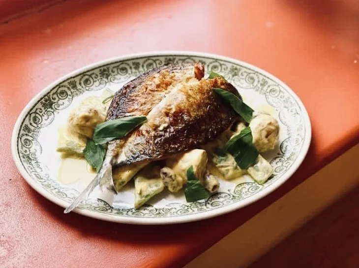 A plated dish featuring a piece of cooked fish with a crispy skin, garnished with green leaves accompanied by a creamy sauce with vegetables on a decorative oval plate.