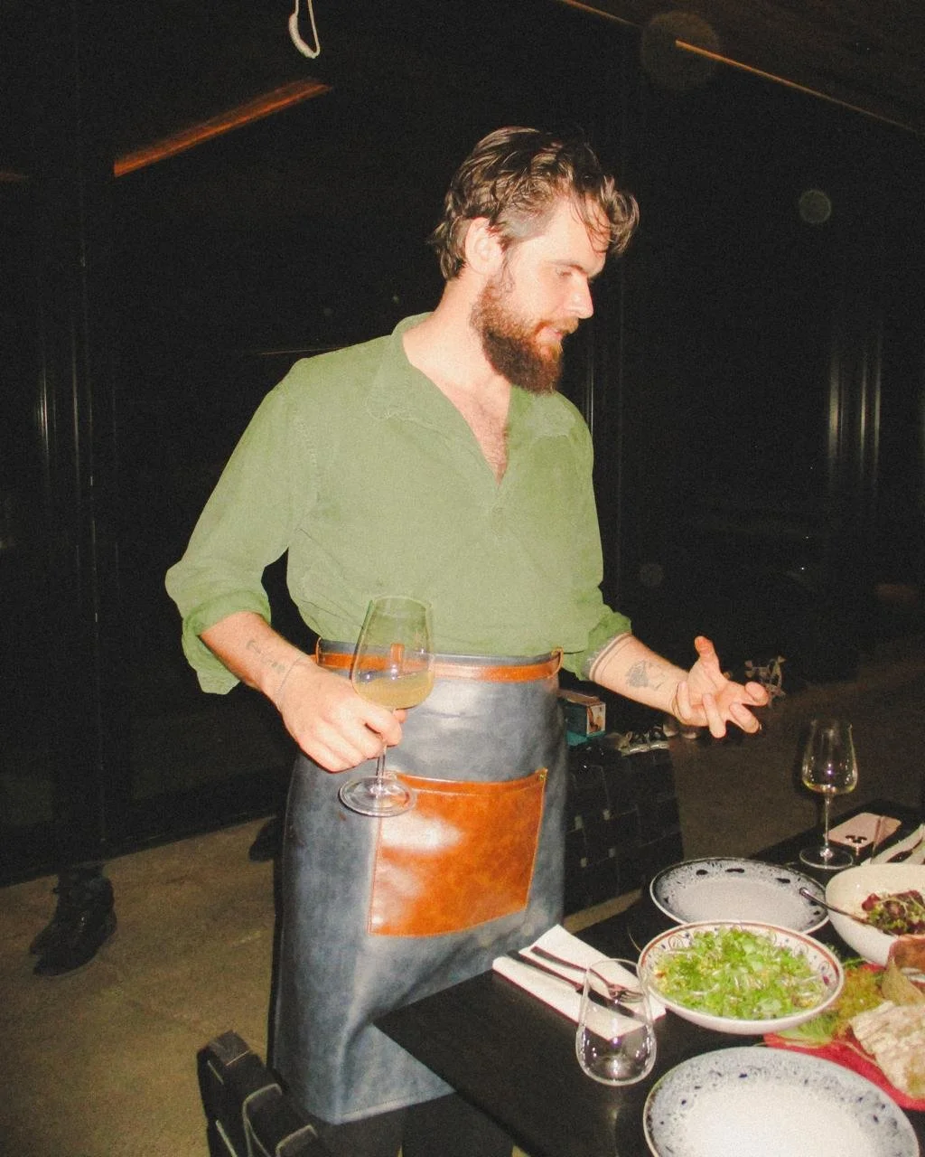 A man with a beard, wearing a green shirt and an apron, is holding a glass of wine or similar beverage and appears to be preparing food or giving instructions at a table with salads and other dishes.