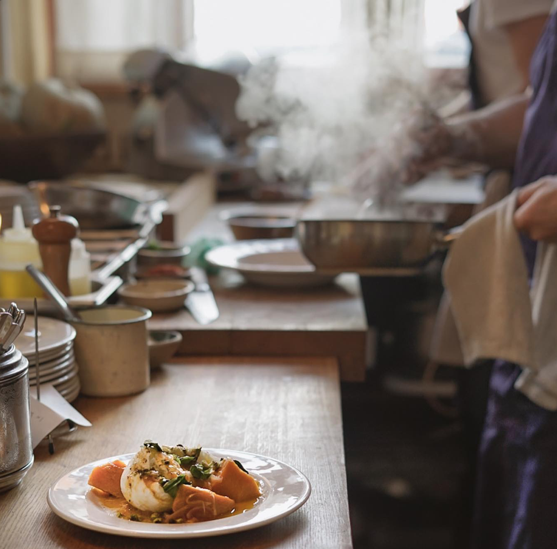 A plate of food, including a poached egg, surrounded by slices of sweet potato or yam, on a wooden kitchen counter. In the background, a person is preparing food with steaming hot dishes, kitchen utensils, and ingredients visible.