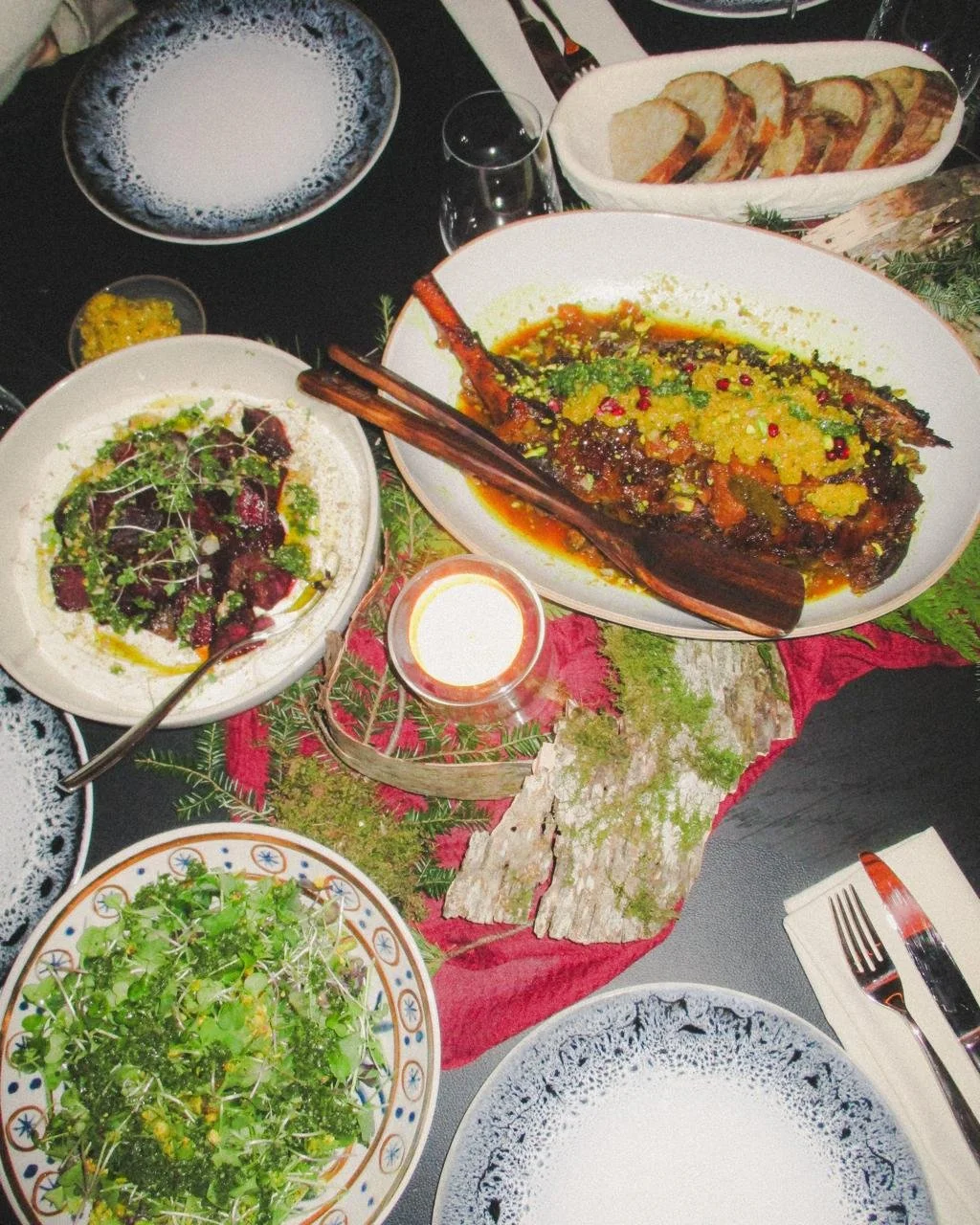 A table with various dishes including a whole cooked fish topped with herbs and sauce, salad with greens and beets, sliced bread, and a bowl of frothy beverage, all decorated with a red cloth, green foliage, and candlelight.