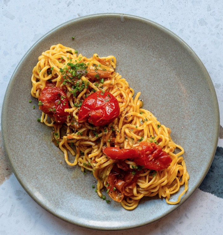 Plate of cooked ramen noodles with cherry tomatoes and green herbs.