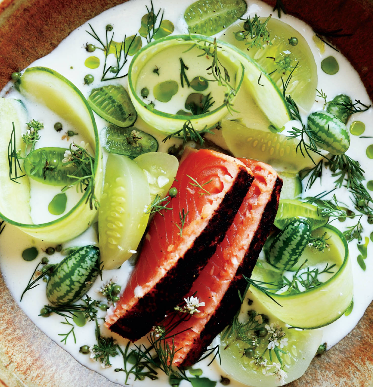 Salmon dish garnished with cucumber slices, herbs, and small watermelon-shaped decorations in a creamy sauce.