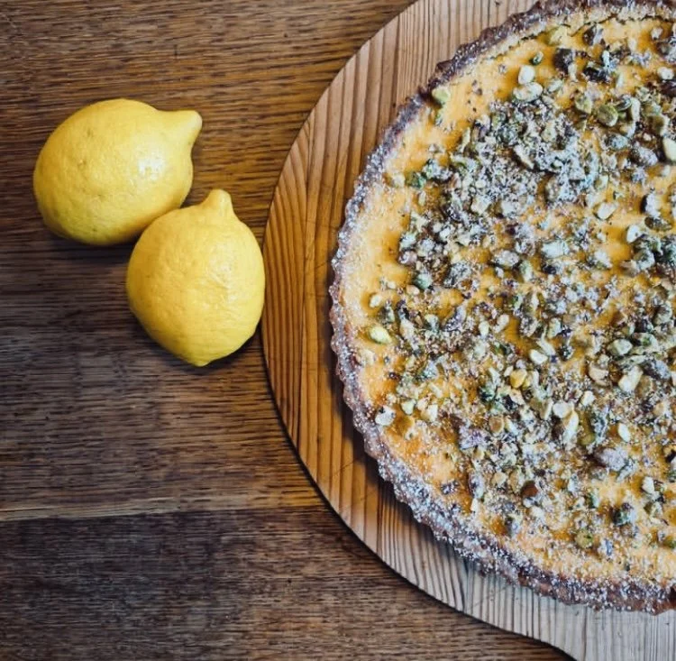 A lemon tart topped with chopped pistachios sits on a wooden cake board next to two fresh lemons on a wooden table.