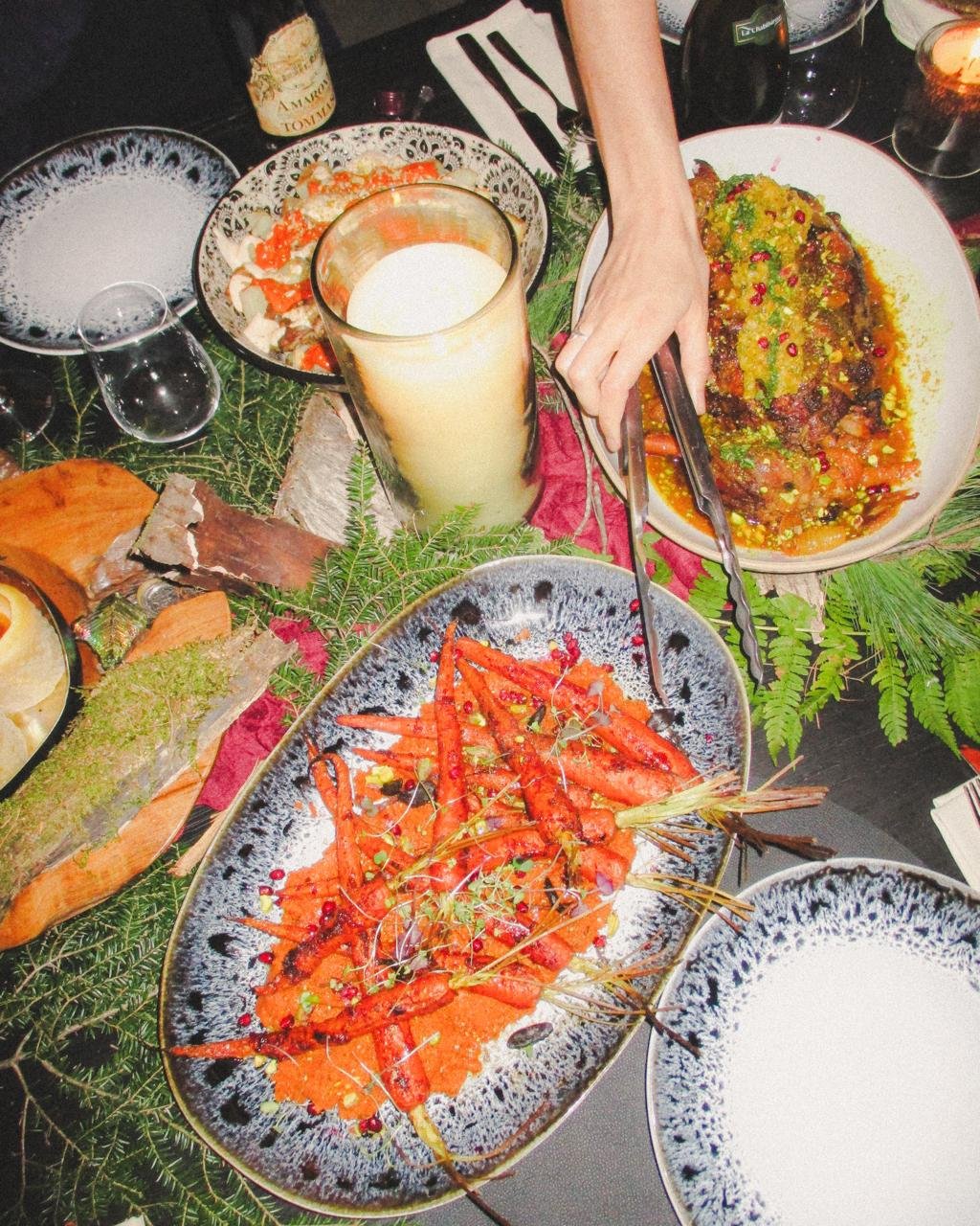 Various dishes on a festive table, including cooked carrots, a large main dish with sauce, a salad, a glass of a creamy beverage, and other drinks, decorated with greenery.