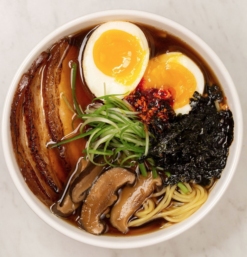 Bowl of ramen noodles topped with sliced pork, soft-boiled egg halves, green onions, mushrooms, shredded nori seaweed, and chili paste in broth.