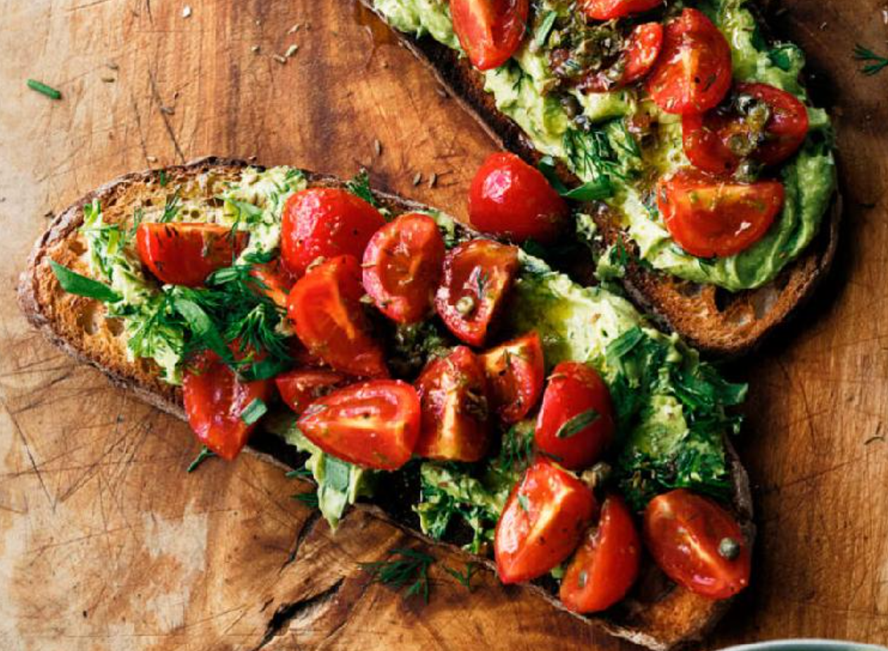 Open-faced toast with avocado, cherry tomatoes, and fresh herbs on a wooden surface.
