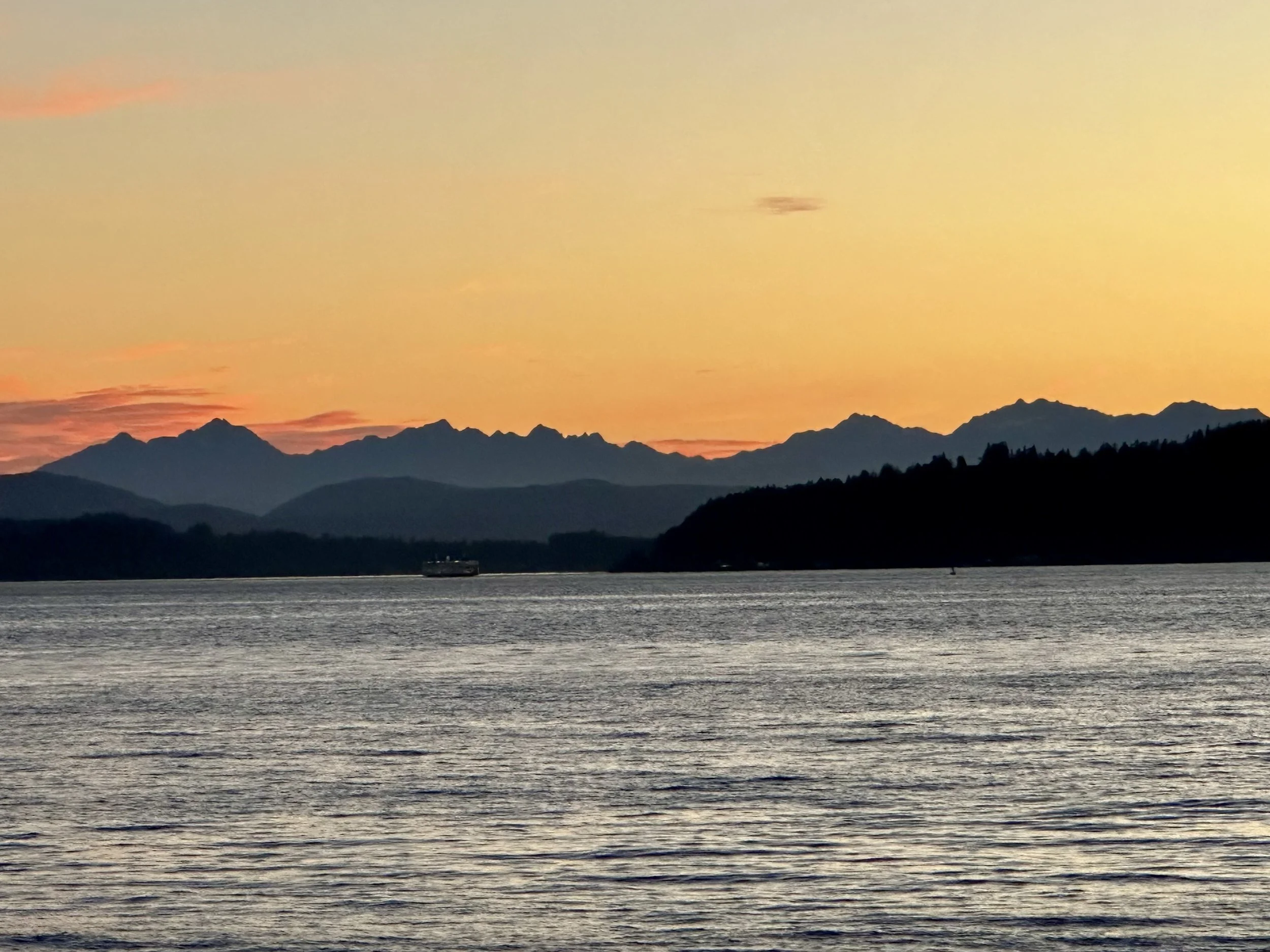 Sunset over a mountainous landscape with a large body of water in the foreground and a boat sailing.