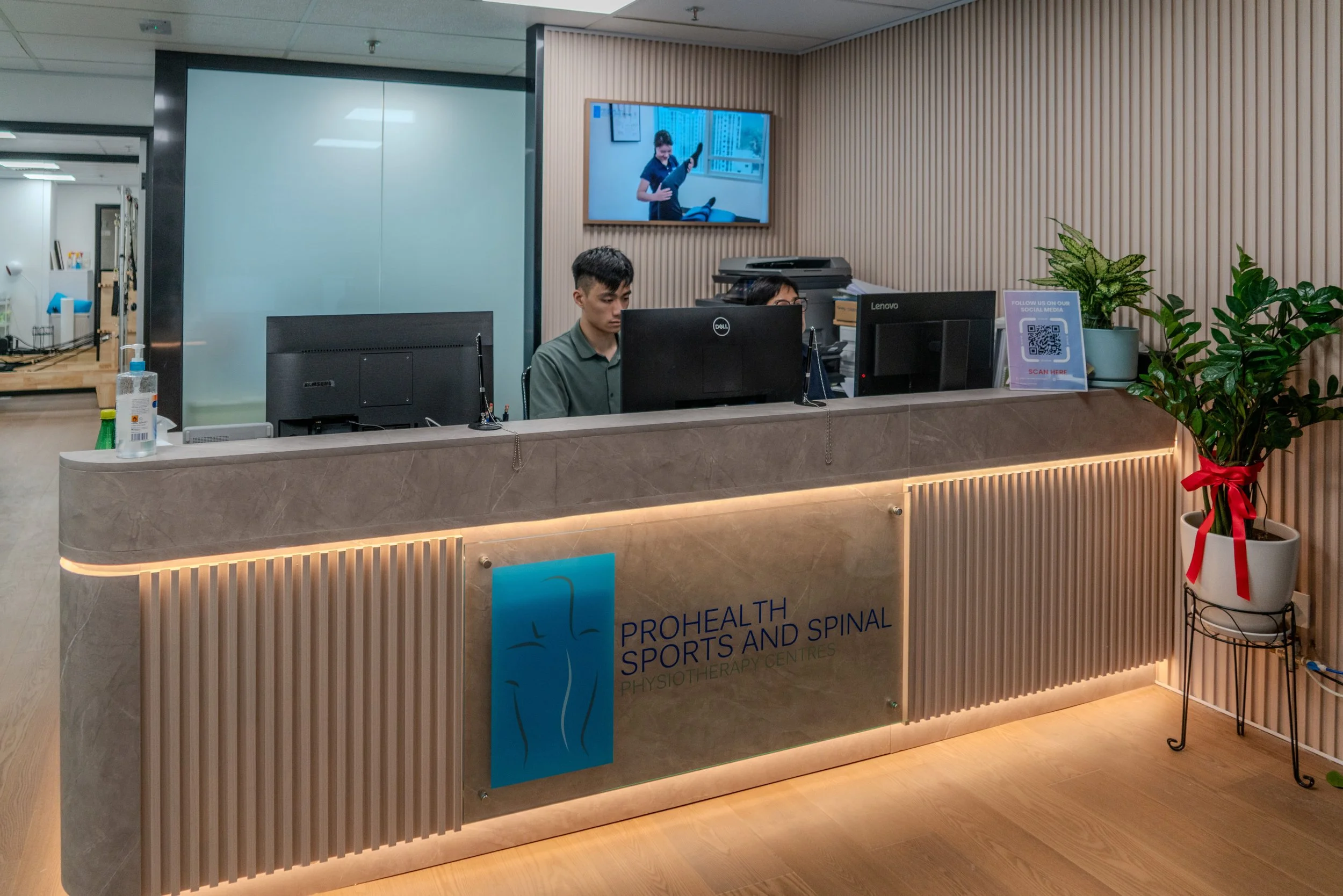 Reception area of Prohealth Sports and Spinal Physiotherapy Centre with informational brochures on the counter, a water cooler, medical posters on the wall, and a person partially visible working at the desk.