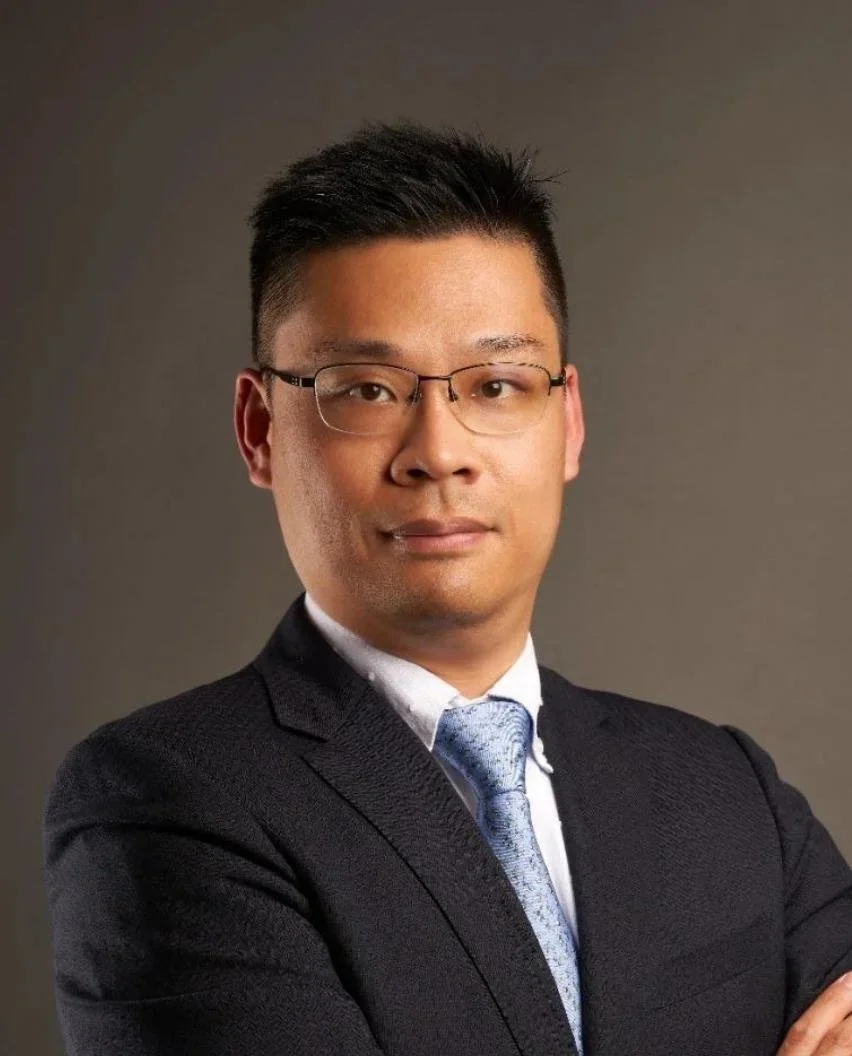 Professional portrait of a man wearing glasses, a dark suit, white shirt, and blue tie, with arms crossed and a neutral expression against a gray background.