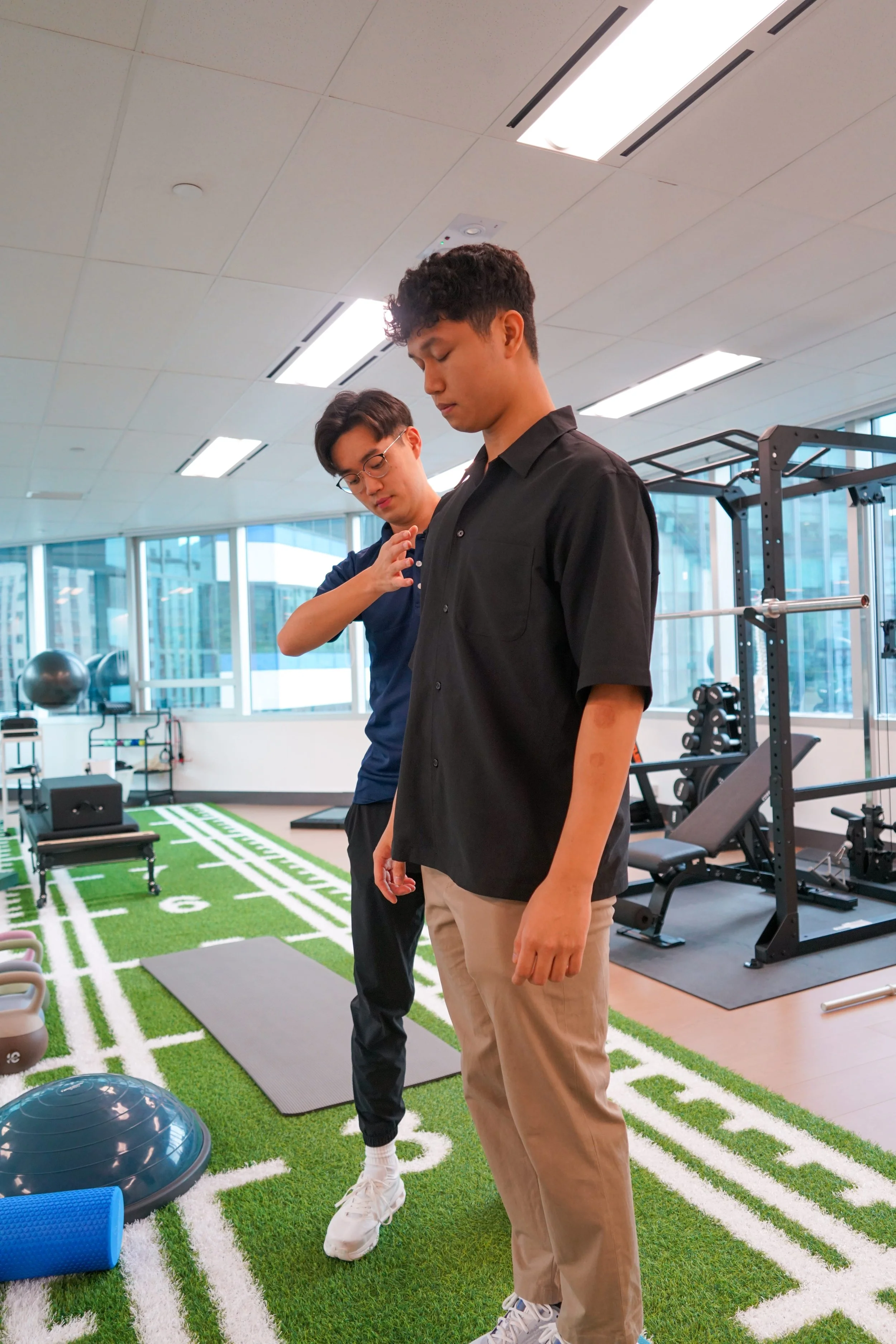 Two young men in a gym. One is standing in front, looking down, wearing a black shirt and beige pants. The other, in a navy shirt and glasses, is behind him, touching his shoulder. Gym equipment and a turf with white lines are visible.