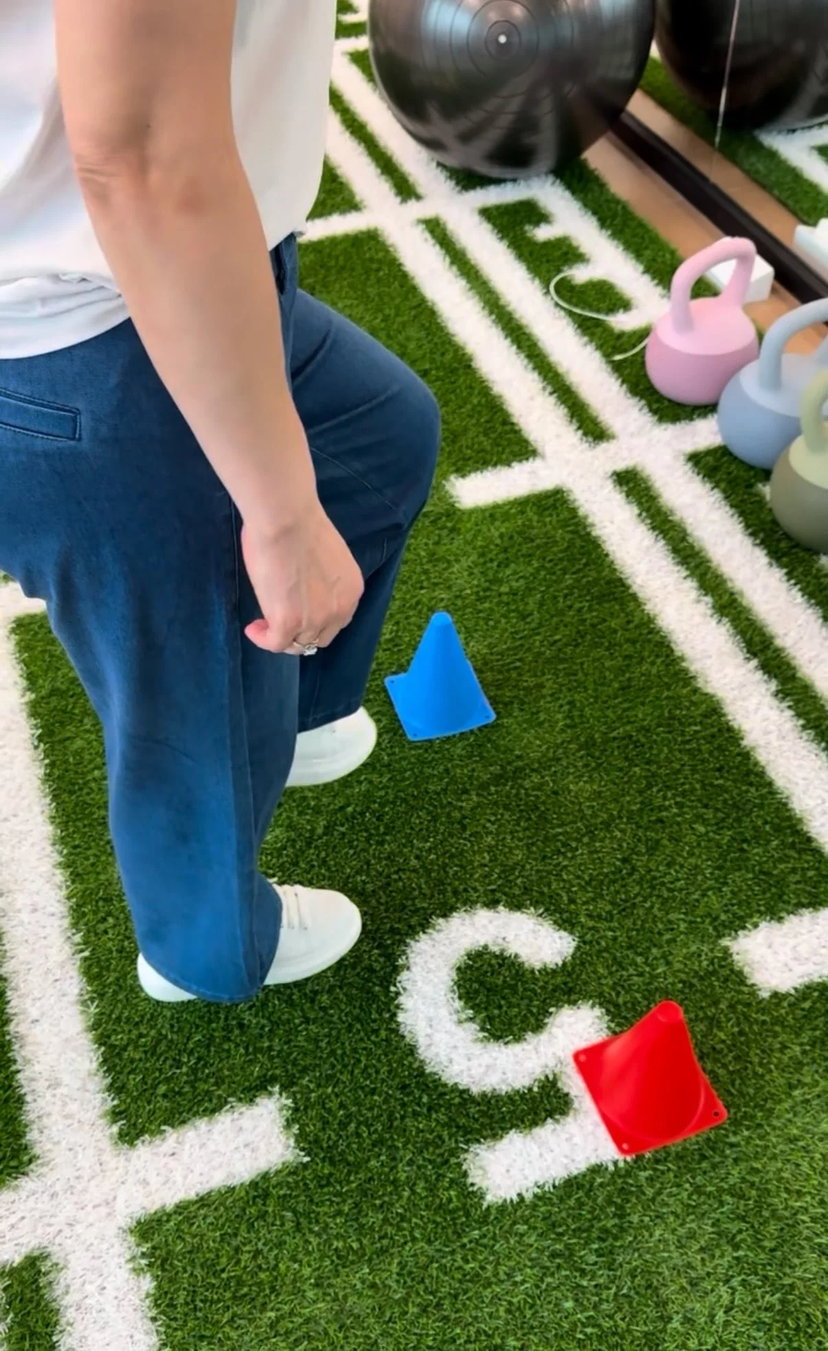 Person in white shirt and blue jeans standing on a green turf with white lines, surrounded by kettlebells and colored plastic cones used for workout or training.