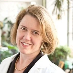Portrait of a woman with blonde hair, wearing a white coat, smiling, in a bright, plant-filled indoor setting.