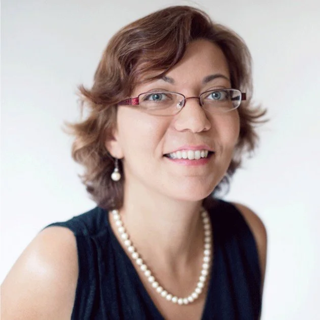 A woman with short brown hair wearing glasses, pearl earrings, and a pearl necklace, smiling at the camera against a plain light background.