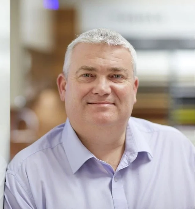 A middle-aged man with short gray hair in a blue button-up shirt, smiling at the camera. The background is blurred with indoor setting elements.