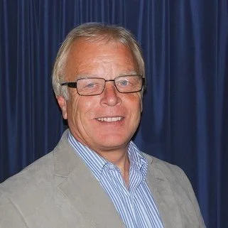 Portrait of a man with glasses, wearing a light gray blazer and a blue striped shirt, standing in front of a blue curtain.