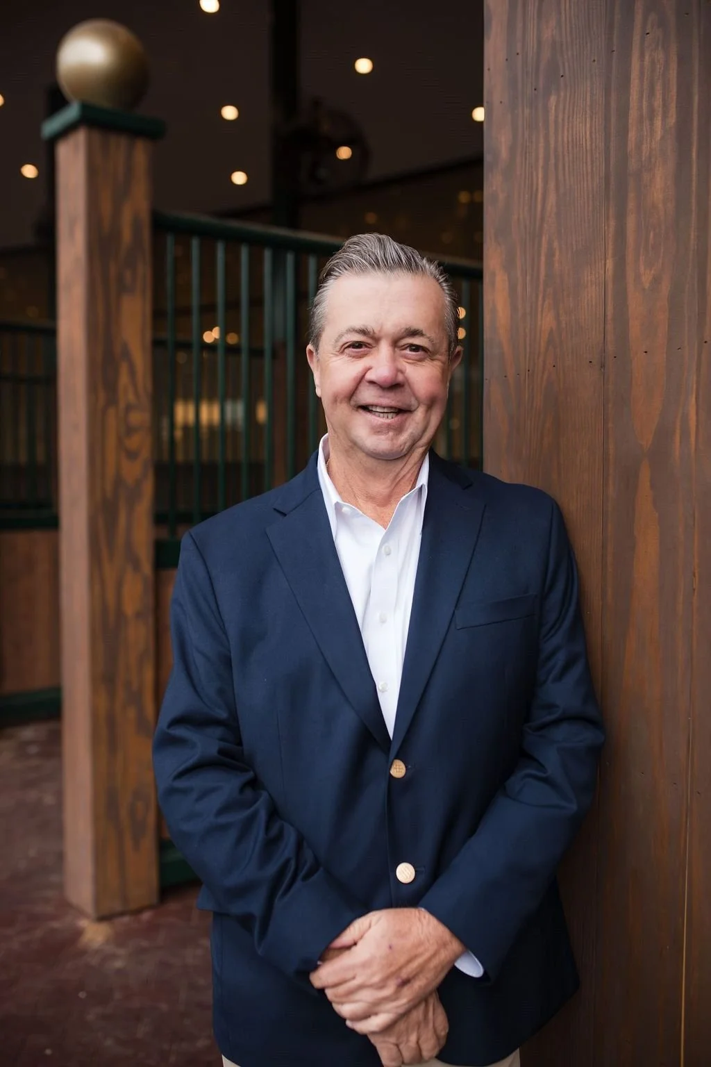 A middle-aged man in a blue blazer and white shirt standing and smiling indoors, next to wooden paneling.