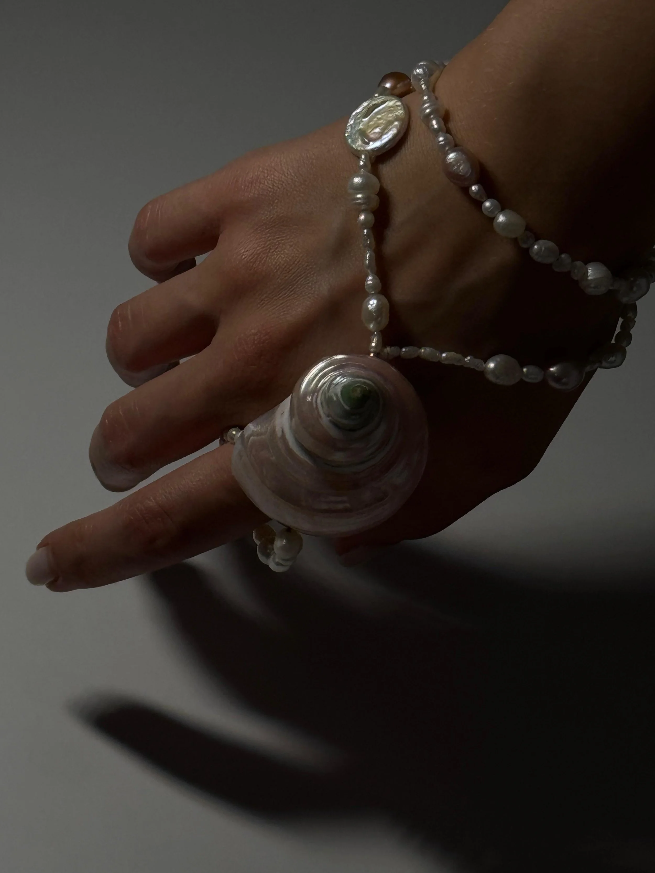 A hand wearing pearl jewelry, including a large pearl shell necklace and matching bracelet against a dark background.