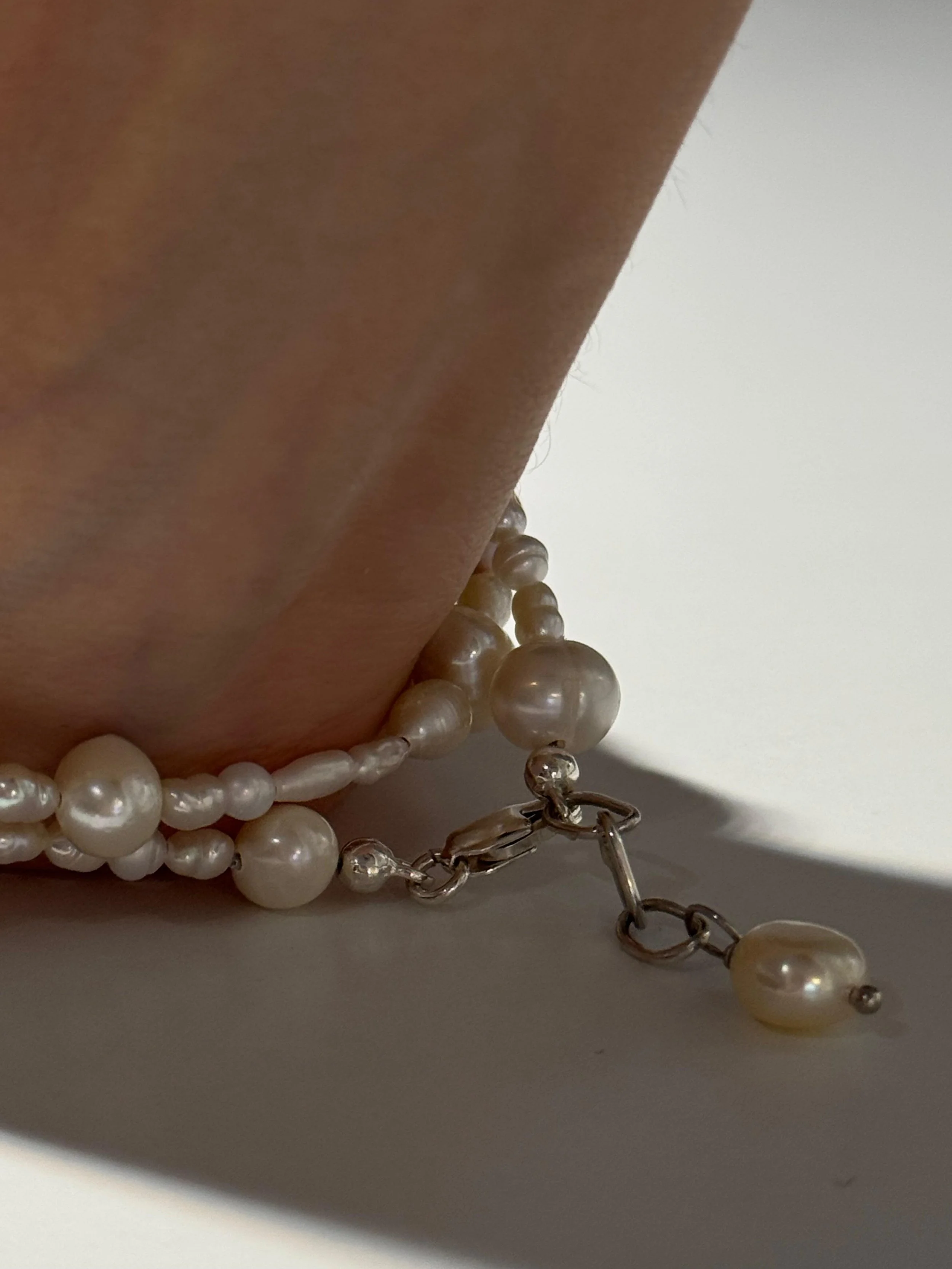 Close-up of a pearl necklace with a silver clasp and dangling pearl charm resting on a white surface, partially shadowed.
