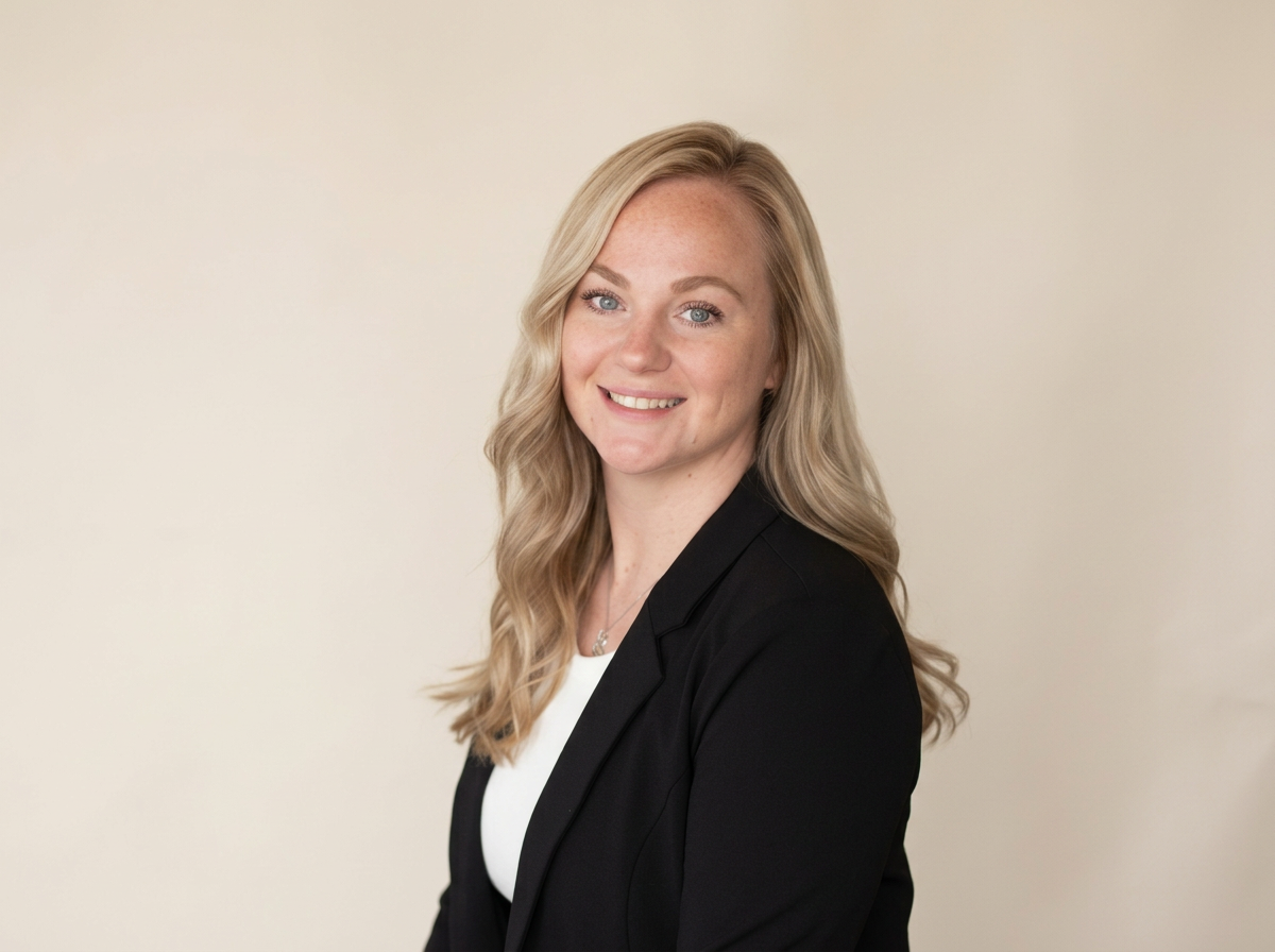 A woman with long blonde hair smiling at the camera, wearing a black blazer over a white top, standing against a plain light-colored background.