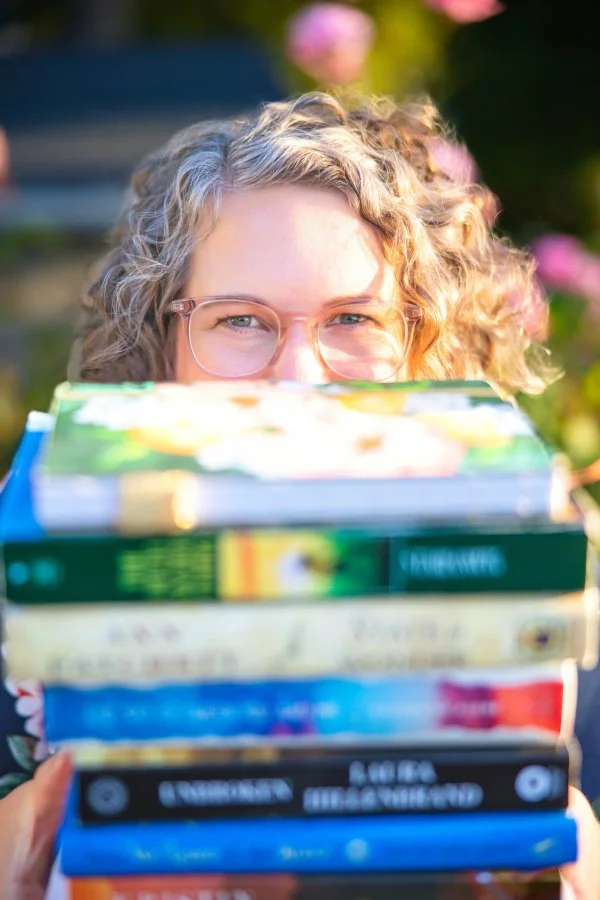 Annie Mesaros peeks playfully over a tall stack of colorful books, her curly hair and pink glasses illuminated by warm sunlight in a garden.