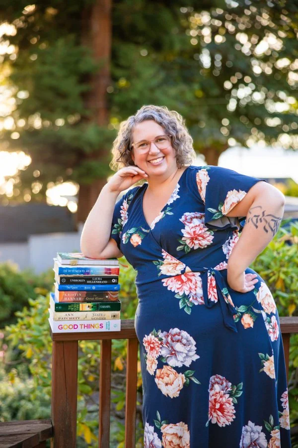 Annie Mesaros smiles on a sunlit wooden deck, wearing a navy blue floral dress and pink glasses, with one hand on her hip and the other resting on a stack of books.