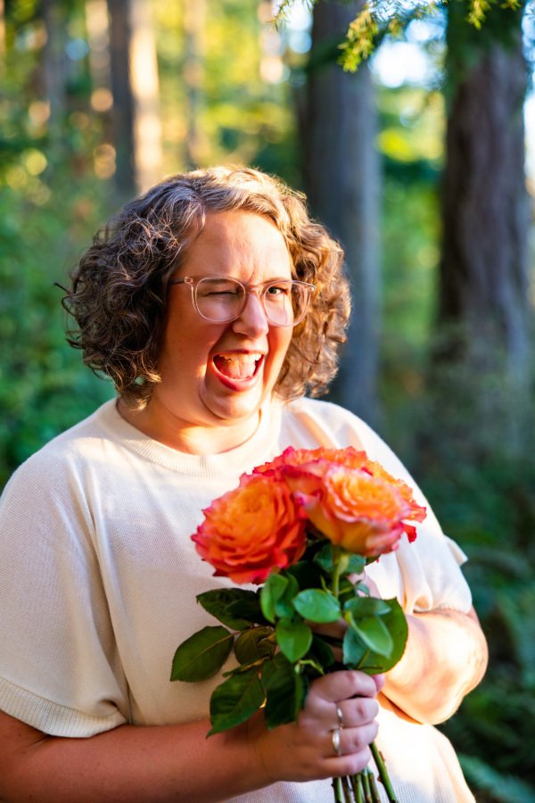 Annie Mesaros smiling and winking while holding a bouquet of roses in the woods.