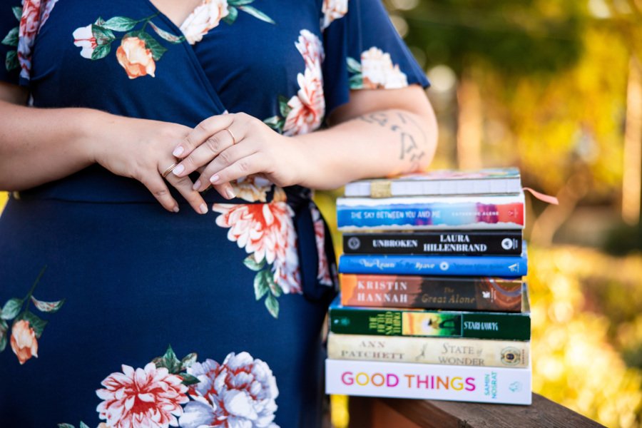 Person in a floral dress holding a stack of books outdoors.