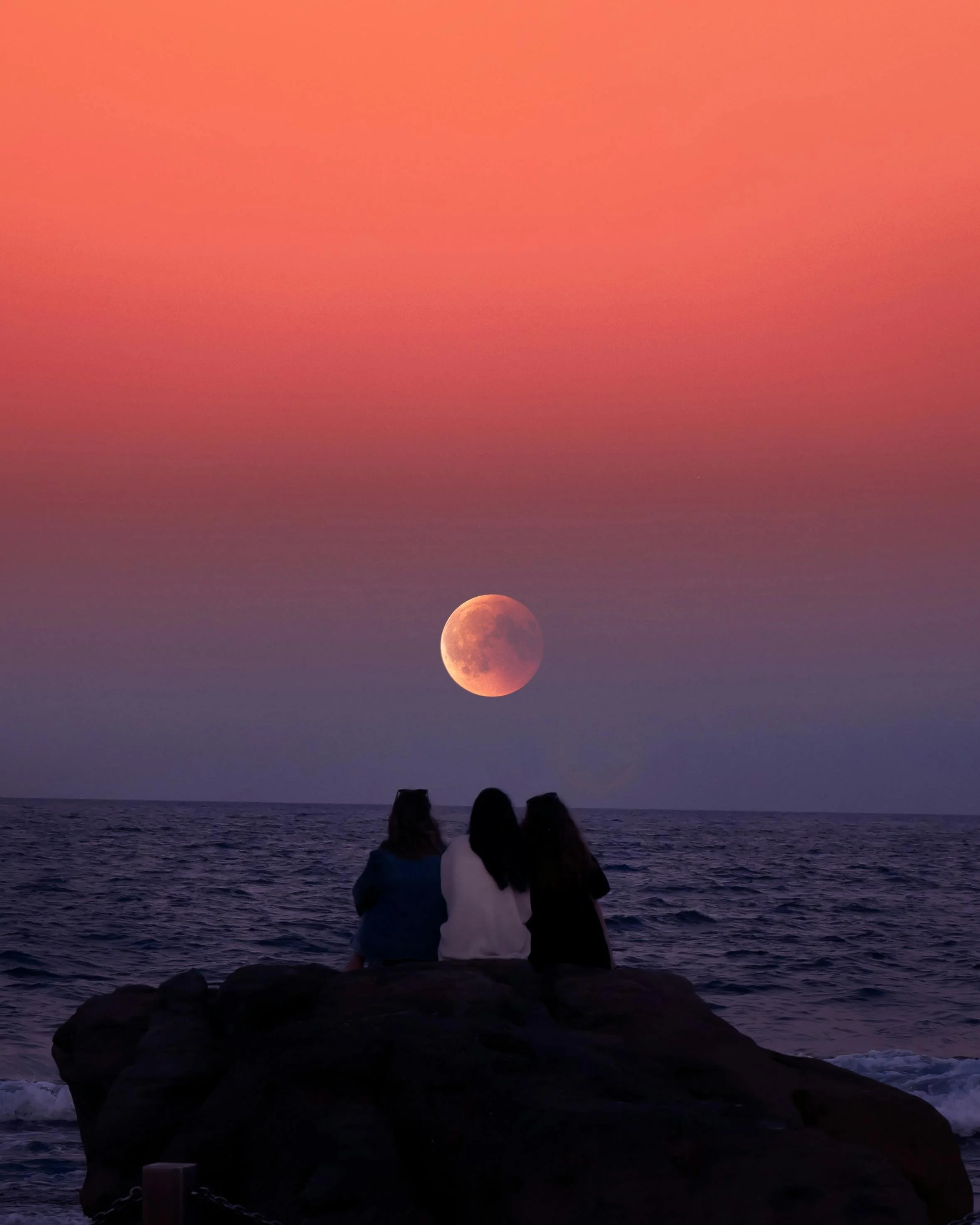 Three women watch a full moon rise over the ocean under a pink sky.