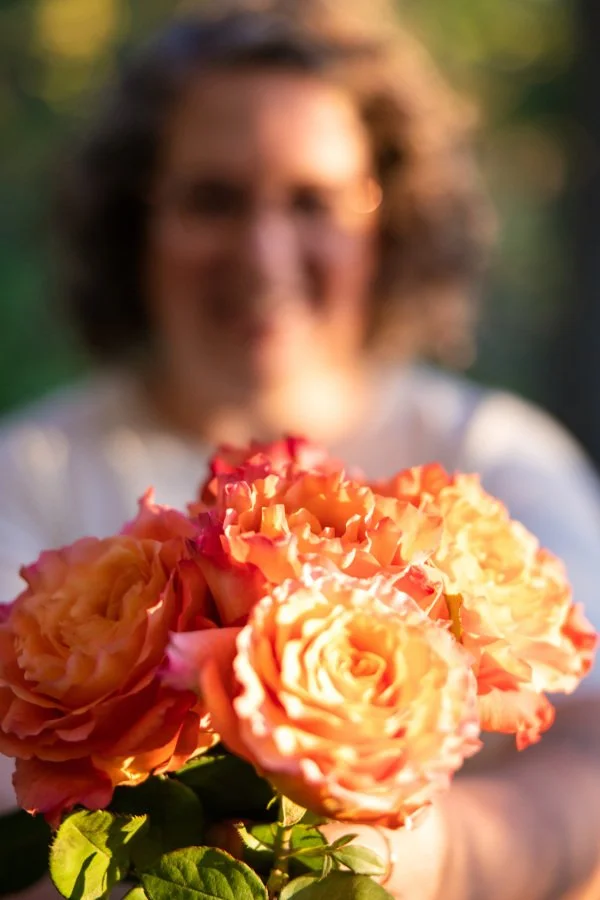 A bouquet of vibrant peach, coral, and golden roses glows in the afternoon sunlight, with a softly blurred person in the background.