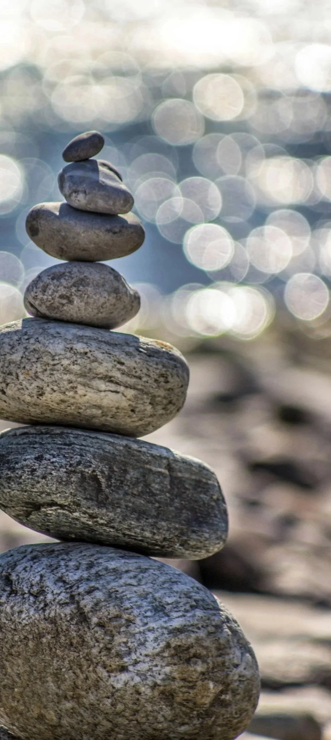 Stacked smooth gray stones on a beach with water in the background and circular bokeh lights.