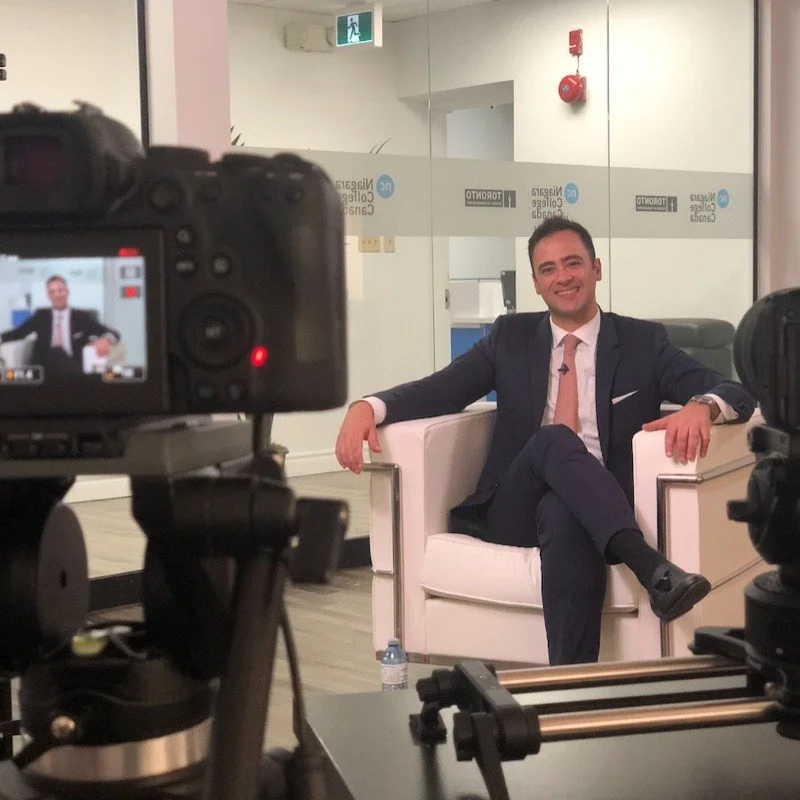 Man sitting on white chair in office setting, being filmed by camera