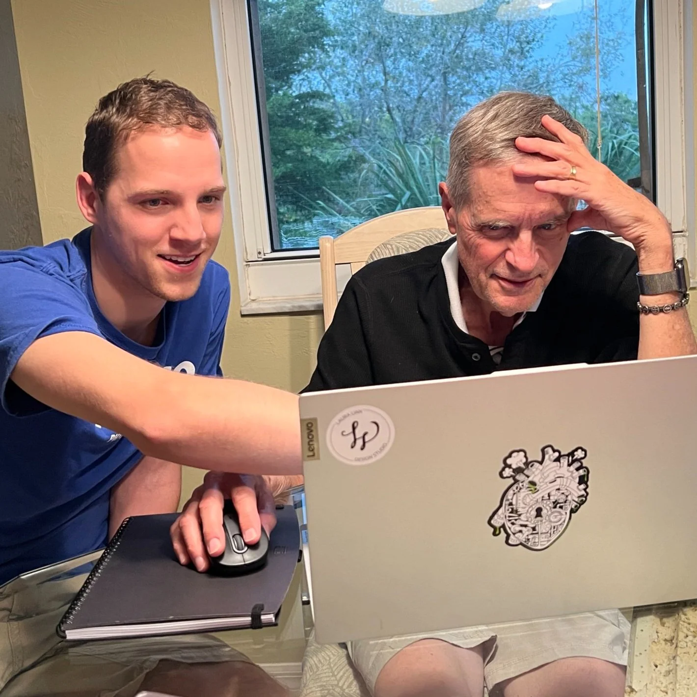 Two people sitting at a table using a laptop together, one man pointing at the screen, the other looking focused with hand on head, a notebook and mouse on the table, window in the background.