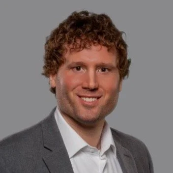 A professional headshot of a person with curly hair, wearing a gray suit and white shirt, smiling against a gray background.