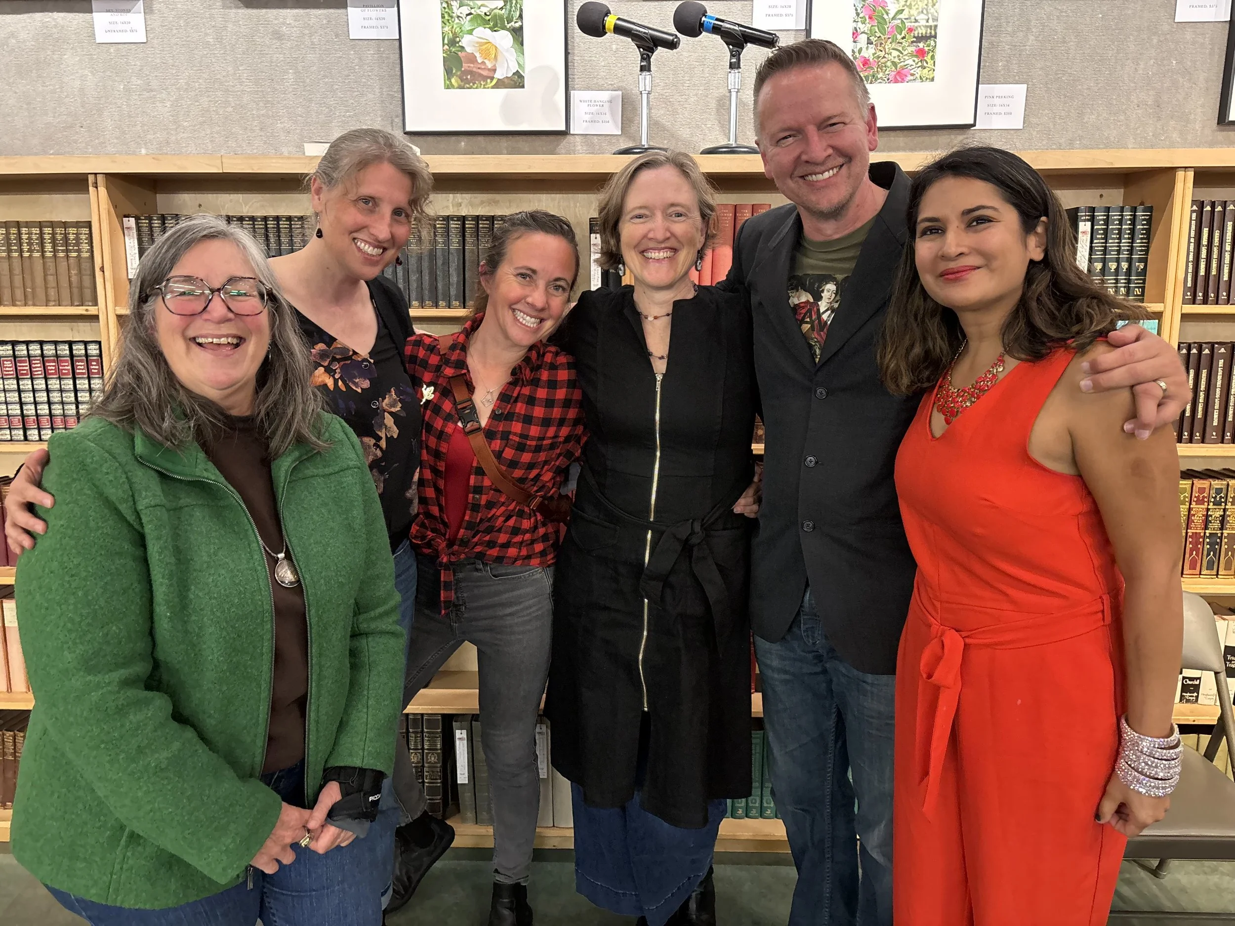 Group of seven people smiling, standing close together in front of a bookshelf with framed floral pictures on the wall behind them.