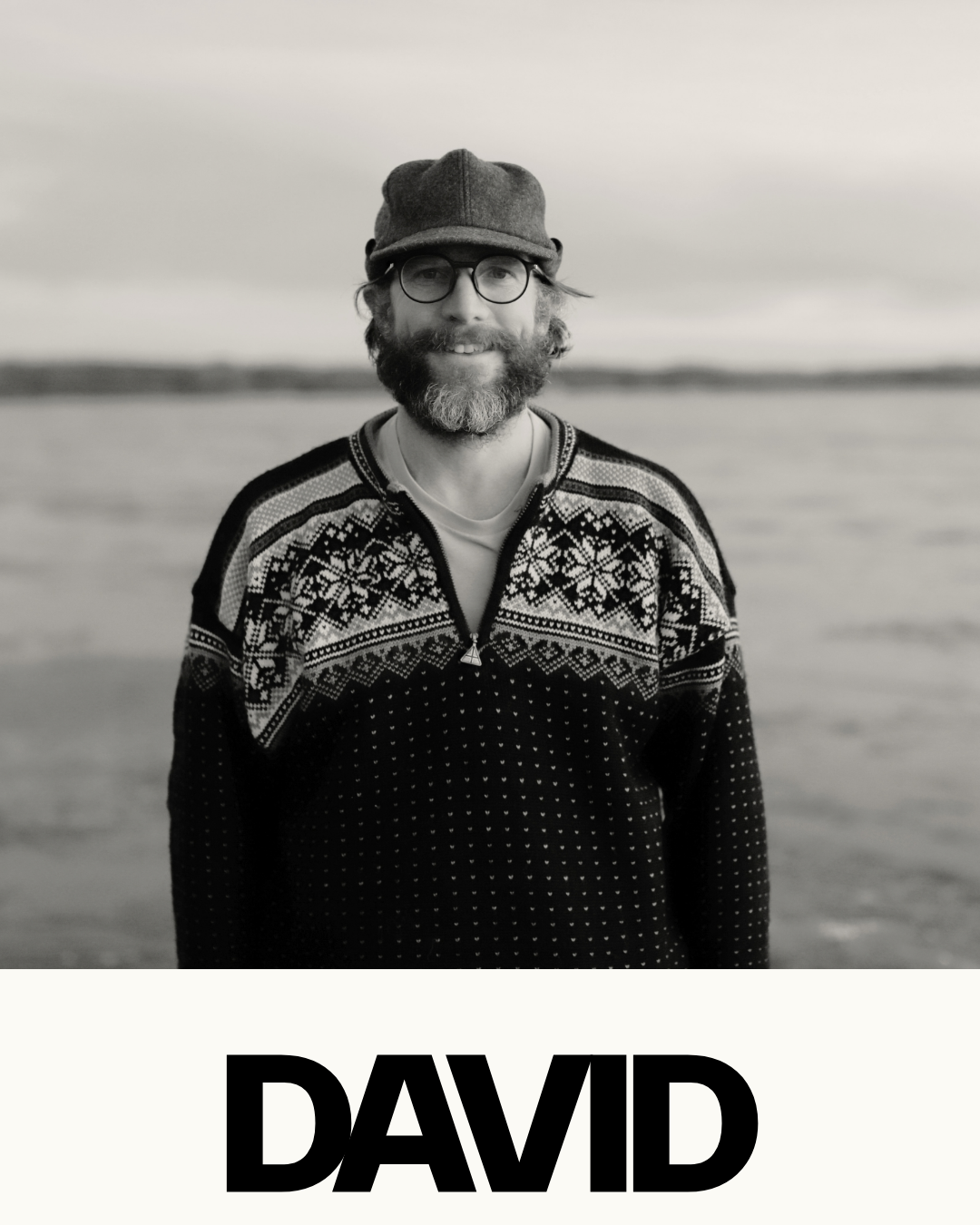 Black and white portrait of a man outdoors, wearing glasses, a beanie, a patterned sweater, and smiling. The background shows water and distant land. The name 'DAVID' is written at the bottom.