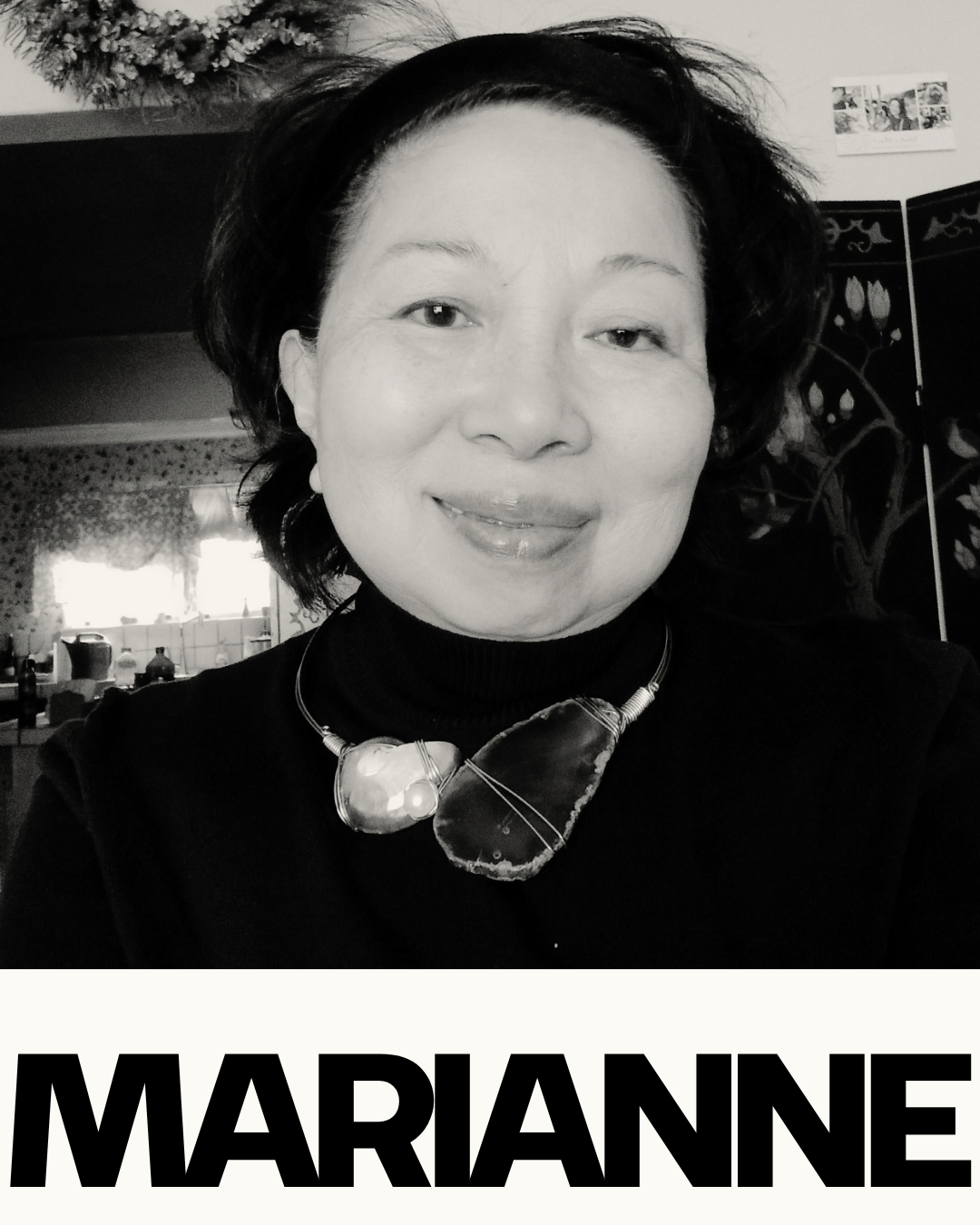 Black and white portrait photo of a woman smiling, wearing a large necklace with a stone pendant. The background shows a kitchen with windows and objects on the counter.