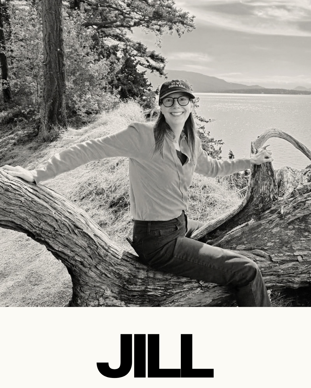 A woman smiling and sitting on a large fallen log near a body of water, with trees and mountains in the background, in a black and white photo.