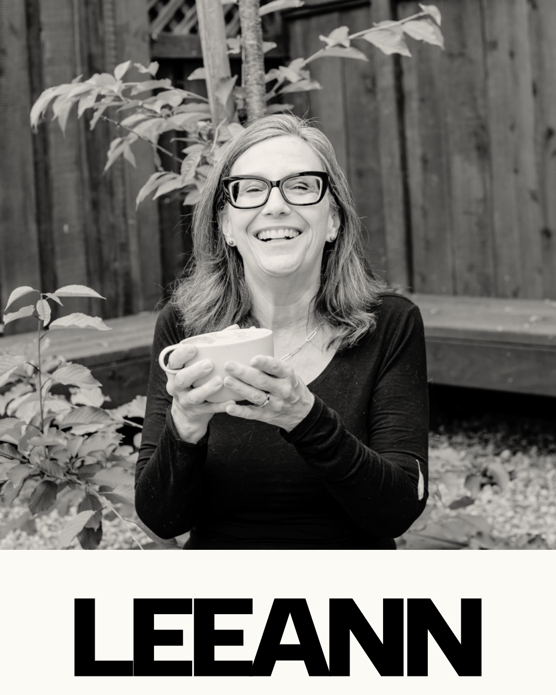 A woman wearing glasses holding a mug and smiling outdoors in a garden, with a wooden fence in the background. The word "LEANN" is written at the bottom.