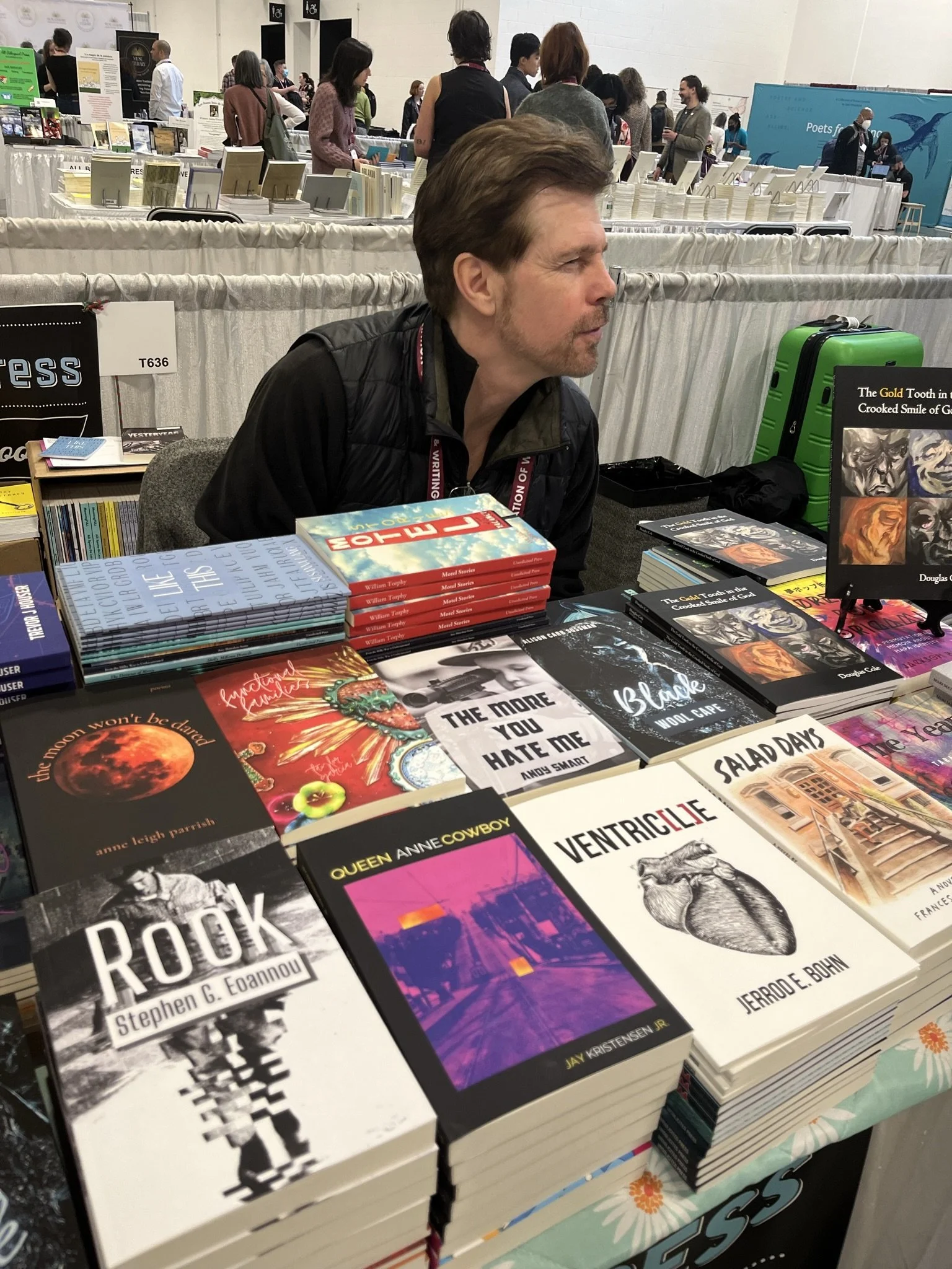 A man with brown hair and facial hair sitting at a table of books at a book fair. The table displays various books, including 'Book,' 'The More You Hate Me,' and other titles. In the background, there are more tables with books and people browsing or conversing.